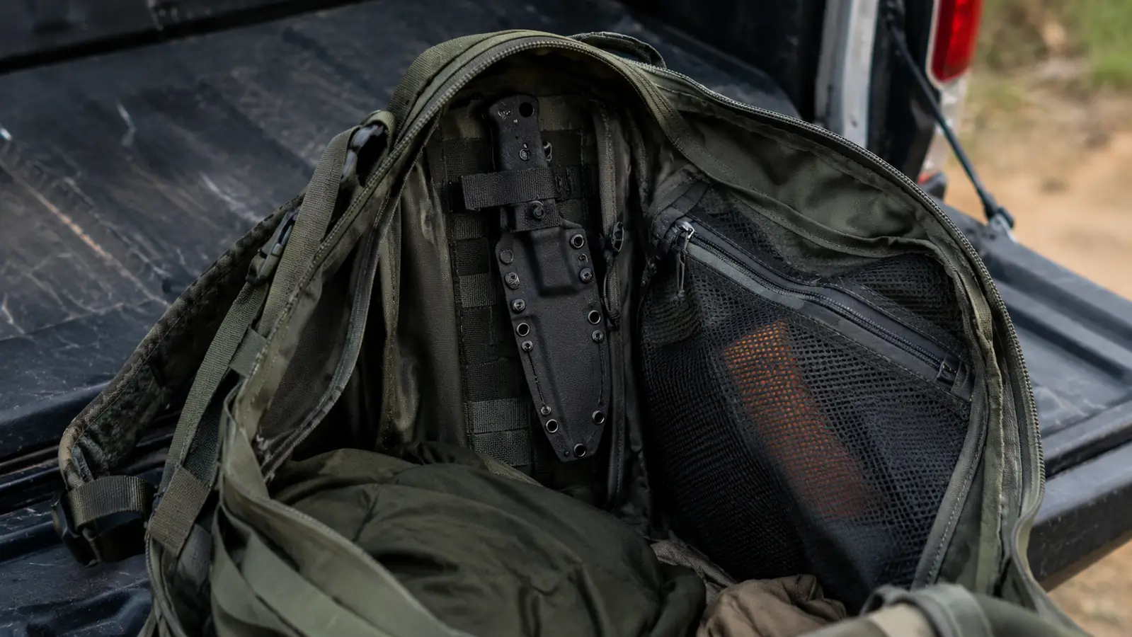 An open backpack on a tailgate with a knife sheath clipped inside the main compartment